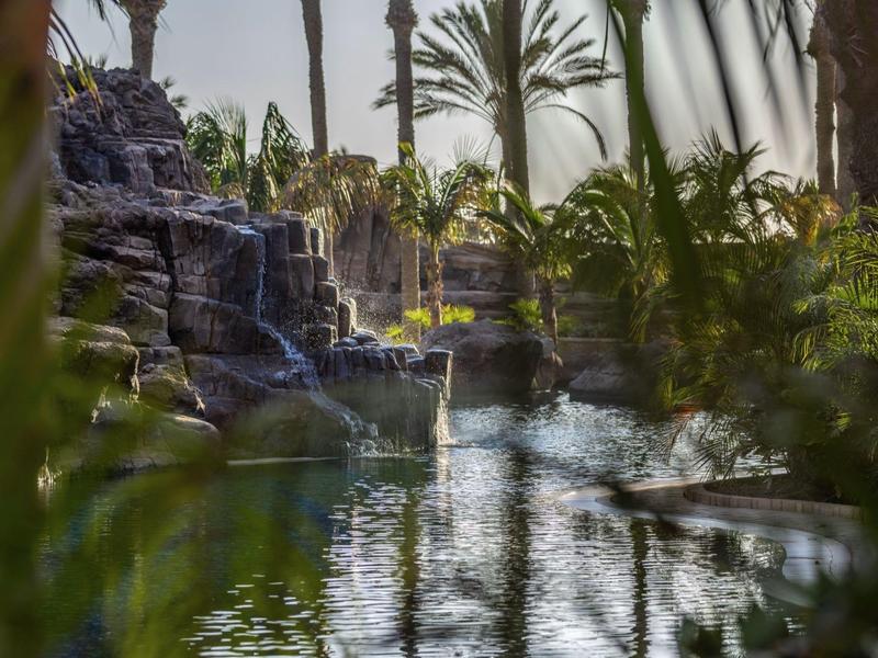 Řeka s vodopádem obklopená palmami a rostlinami v tropické zahradě hotelu.