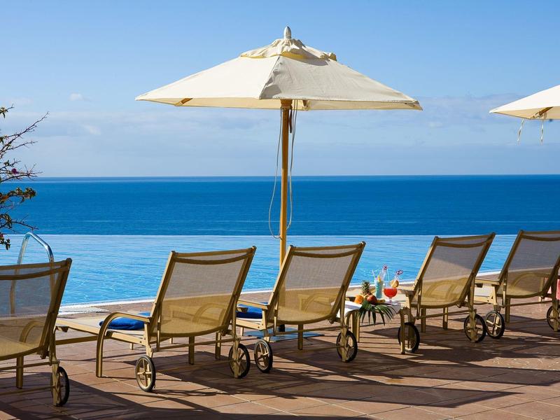 Plážová křesla a slunečníky na dřevěné terase s výhledem na klidné modré moře pod jasnou oblohou.