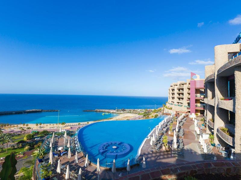 Nekonečný bazén s výhledem na oceán a velkými budovami letoviska pod jasnou modrou oblohou.