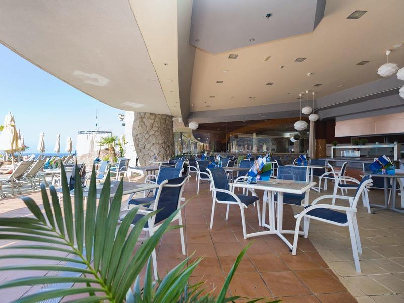 Venkovní posezení hotelové restaurace s bílými stolem a modrými židlemi pod střechou.