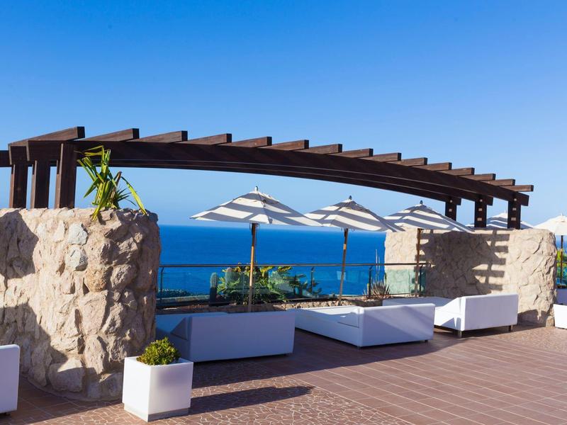 Terasa s výhledem na oceán, pergolou, lehátky a slunečníky pod jasnou modrou oblohou.