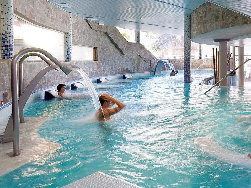 Vnitřní bazén s vodními tryskami a lidmi odpočívajícími pod kamennými zdmi a okny