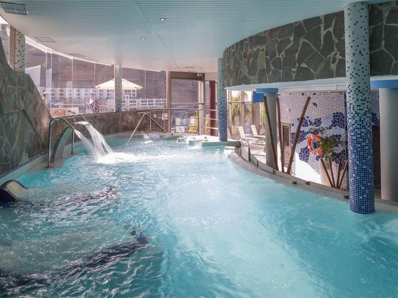 Vnitřní bazén hotelu s kamennými zdmi, vodními tryskami a velkými okny pro přirozené světlo.