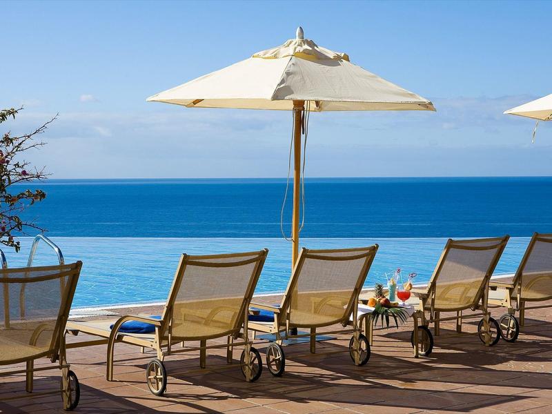 Lehátka a slunečníky na dřevěné terase s výhledem na klidný modrý oceán za slunečného dne.
