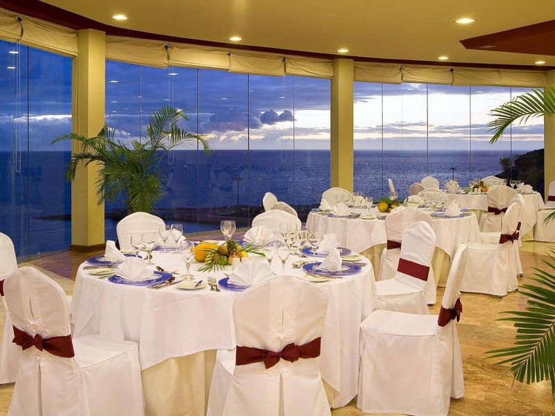 Elegantní jídelna s kulatými stoly, bílými židlemi a výhledem na oceán přes velká okna.