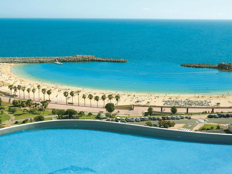 Pohled na pláž s palmami a bazénem v popředí pod jasnou modrou oblohou.