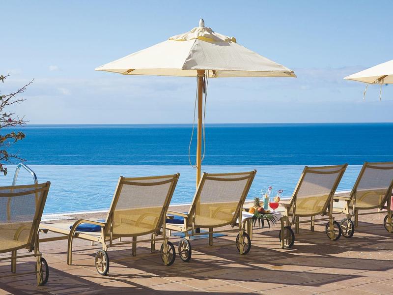 Lehátka a slunečníky na terase s výhledem na klidné moře pod jasnou oblohou.