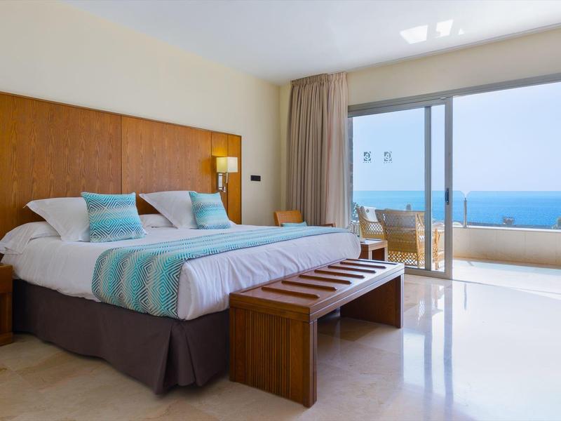Prostorný hotelový pokoj s manželskou postelí, dřevěným nábytkem a balkonem s výhledem na oceán.