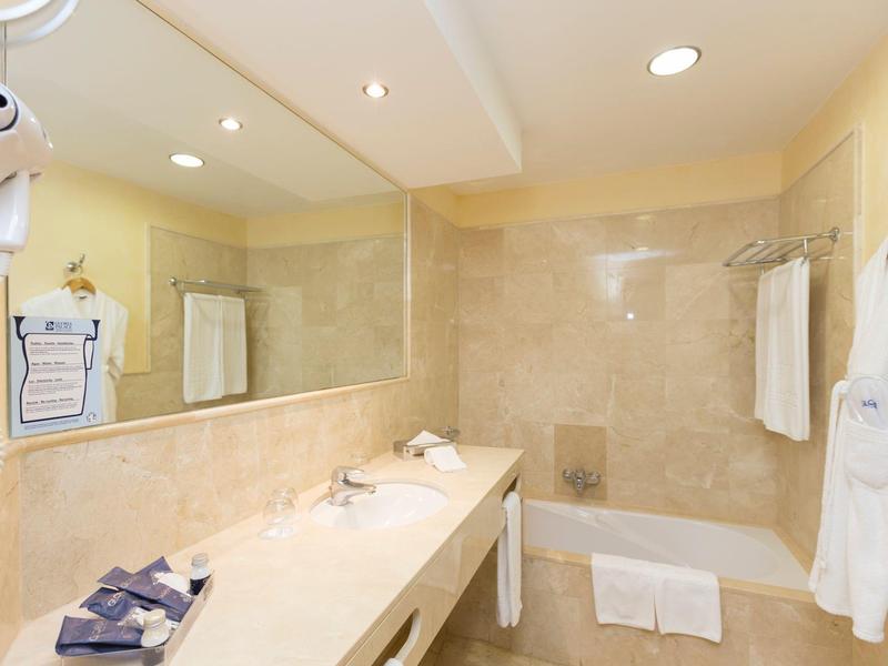 Světlá koupelna s béžovými dlaždicemi, vanou, dlouhým umyvadlem, ručníky a županem.