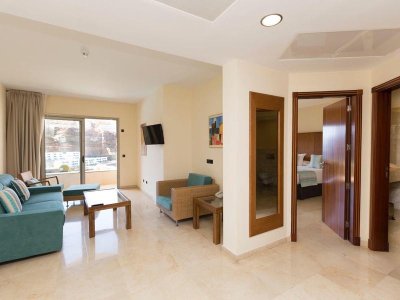 Světle hotelový pokoj s modrozelenou pohovkou, stolkem, televizí a otevřenými dveřmi do koupelny.