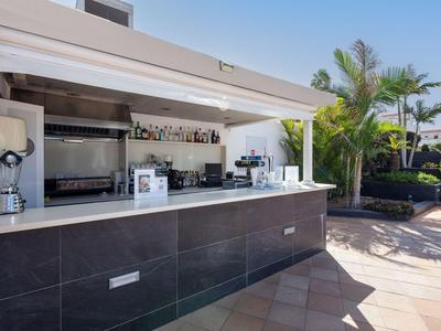 Moderní venkovní bar s židlemi, kuchyní a palmami za slunečného počasí.