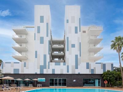 Moderní hotel s bílými věžemi, řadami balkonů a bazénem v popředí pod modrou oblohou.