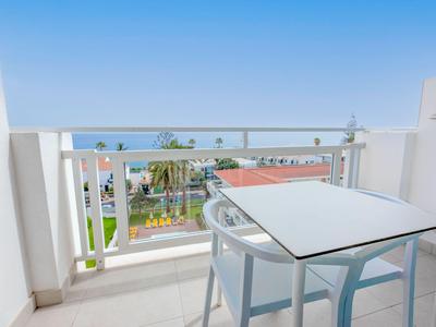Balkon s proskleným zábradlím a bílým stolem se židlemi, výhled na zahradu a budovy pod jasnou oblohou.