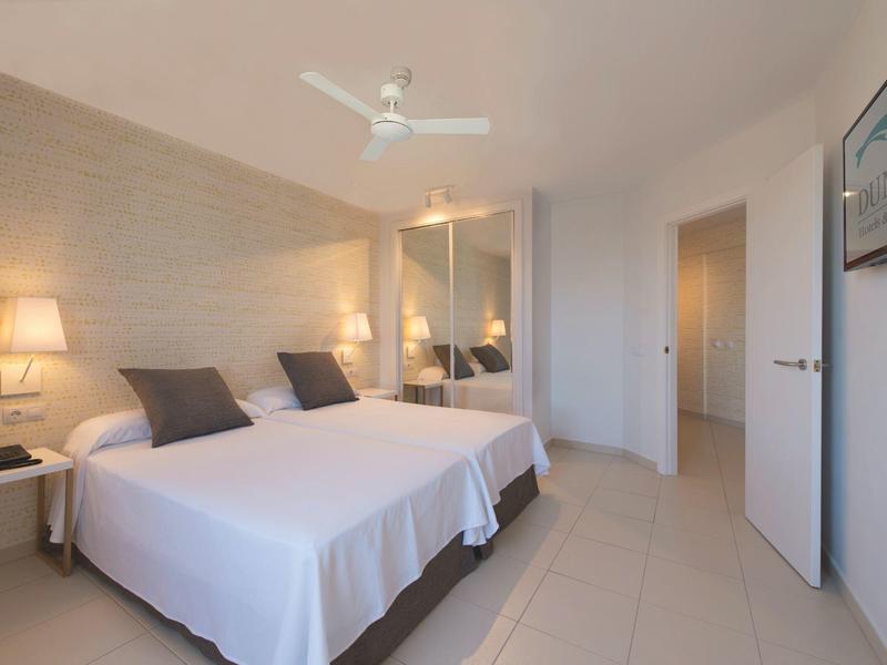 Chambre d'hôtel lumineuse avec deux lits, ventilateur de plafond et décor minimaliste.