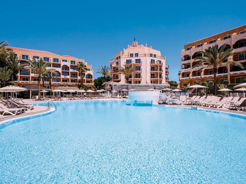 Grande piscine d'hôtel avec chaises longues et palmiers sous un ciel dégagé.