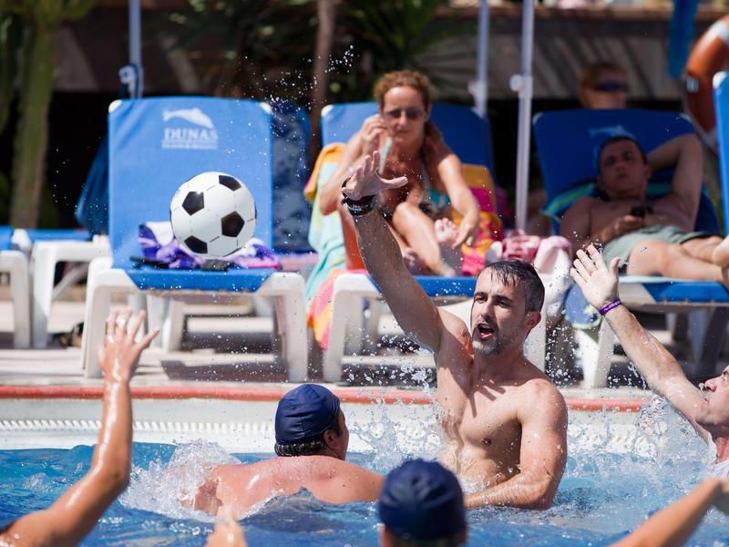 Plusieurs hommes jouent au water-polo dans une piscine avec des personnes sur des chaises longues bleues en arrière-plan.
