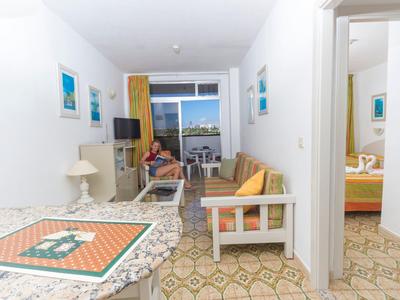 Helles Wohnzimmer mit bunter Sofagarnitur, Teppich und Blick auf Balkon mit Meer. Schlafzimmer rechts sichtbar.
