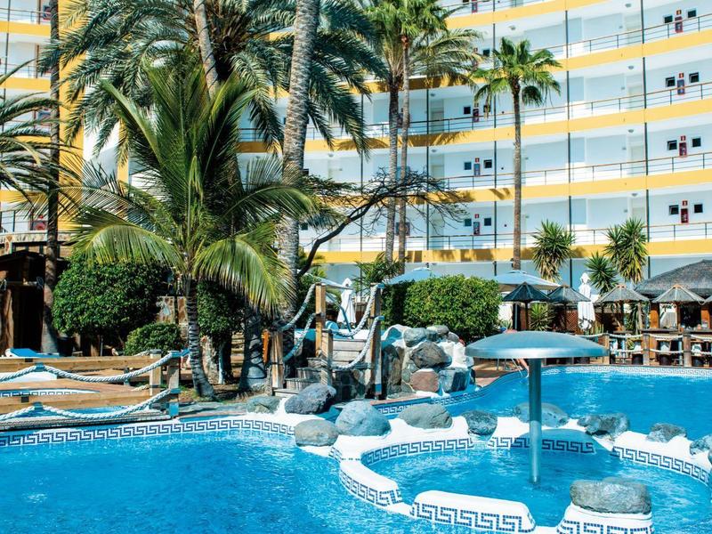 Hôtel avec grande piscine entourée de palmiers et parasols sous un ciel bleu.