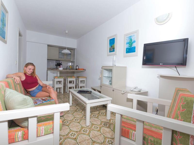 Salon lumineux avec mobilier rétro, canapé, TV murale et cuisine ouverte en arrière-plan.