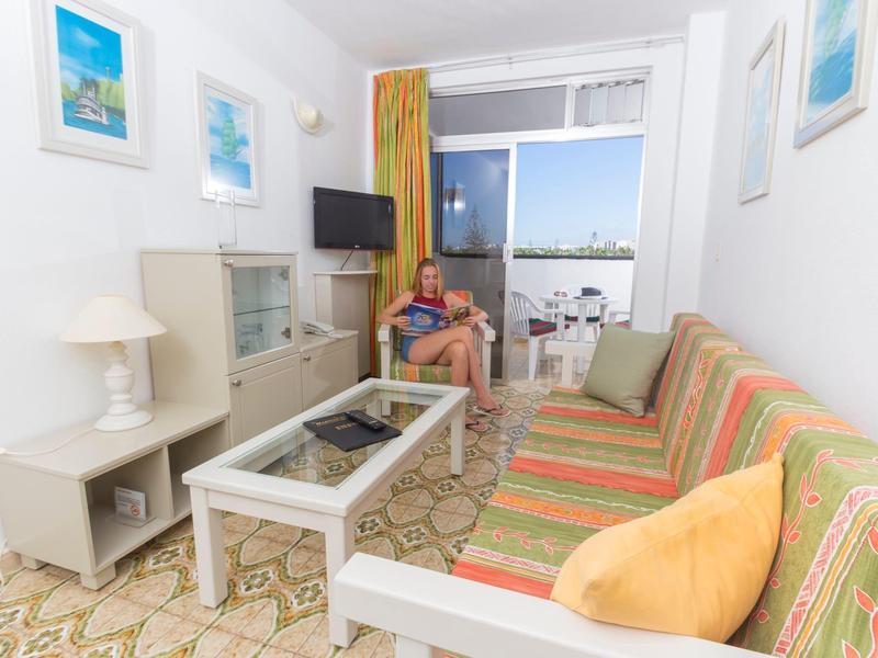 Salon lumineux avec canapé coloré, table en verre et balcon ouvert avec vue sur la mer.