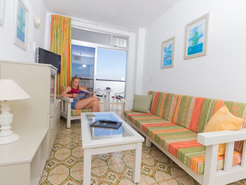 Salon lumineux avec canapé coloré, table blanche et vue sur la mer à travers la porte vitrée.