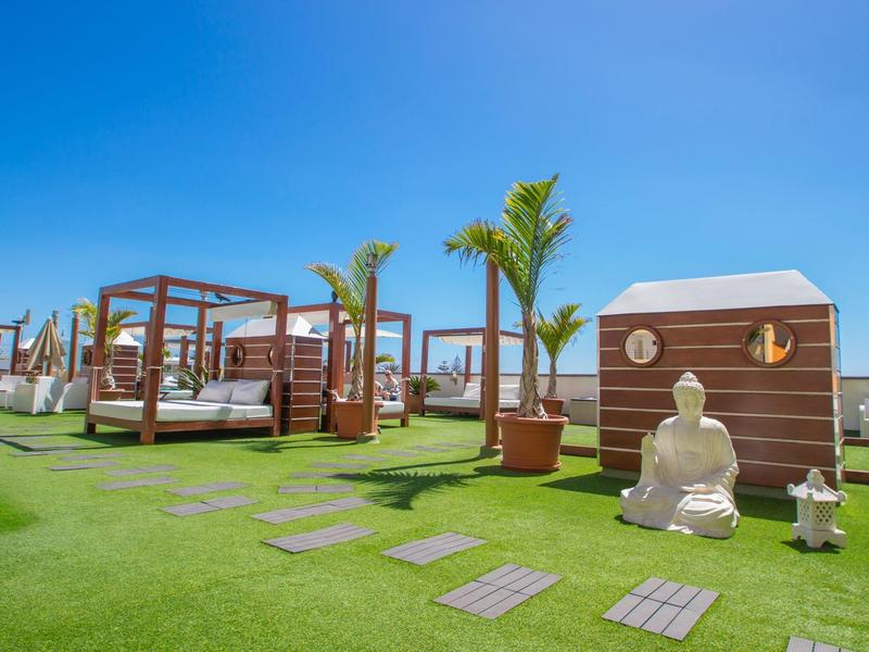 Espace extérieur luxueux avec pelouse verte, pavillons, palmiers et statue de Bouddha sous un ciel bleu clair.