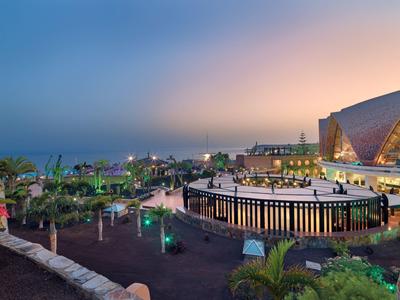 Landschaft mit moderner Architektur, beleuchteten Wegen, Pflanzen und Meer am Abend.