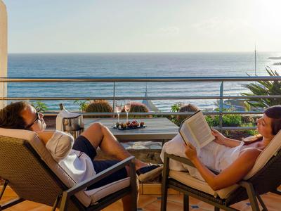 Zwei Personen entspannen auf Balkonliegen mit Meerblick, eine liest, die andere sitzt entspannt.