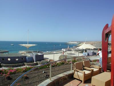 Küstenpromenade mit Sitzgelegenheiten, Blumenbeeten und Blick aufs blaue Meer bei klarem Himmel.