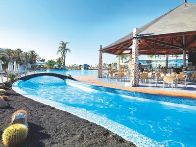 Moderner Außenpool mit blauem Wasser, Brücke, Palmen und überdachtem Sitzbereich bei klarem Himmel.