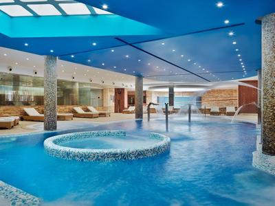 Moderner Innenpool mit rundem Whirlpool, Säulen und blauer Decke, umgeben von Liegestühlen.