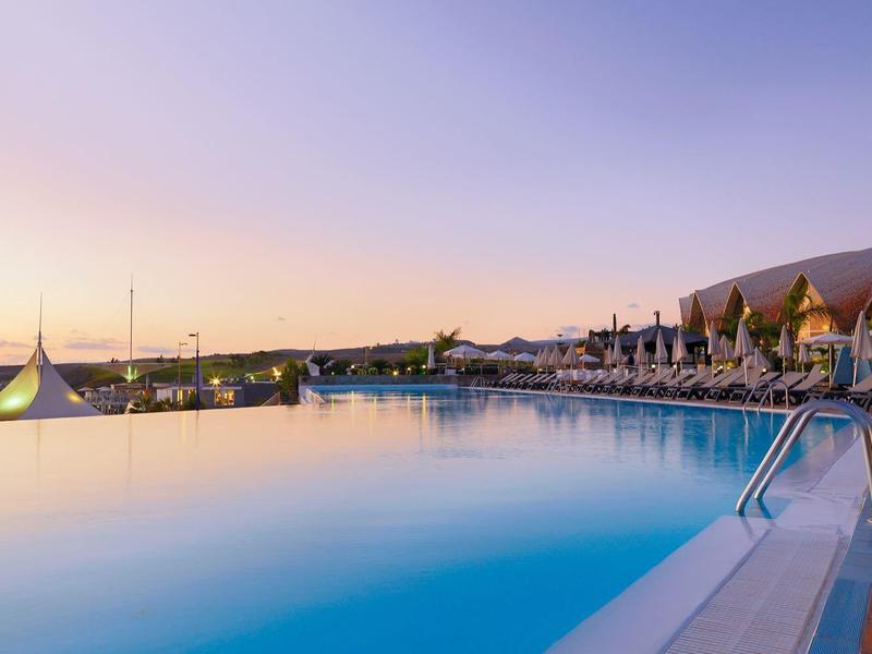 Ein großer, ruhiger Pool mit klarem Wasser und einer Terrasse mit Liegestühlen unter Palmen bei Sonnenuntergang.