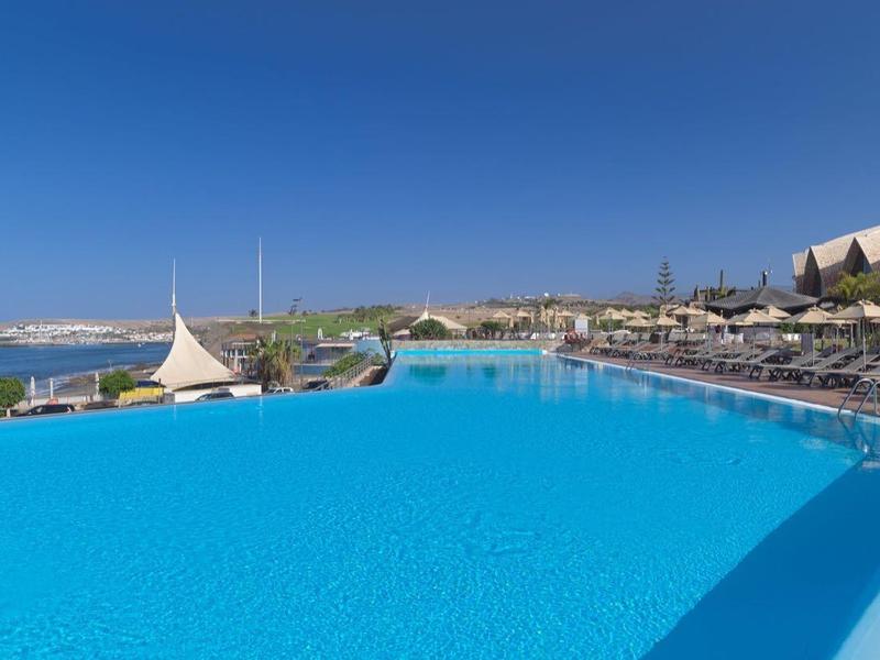 Großer blauer Infinity-Pool mit Liegestühlen und Meerblick unter klarem Himmel.