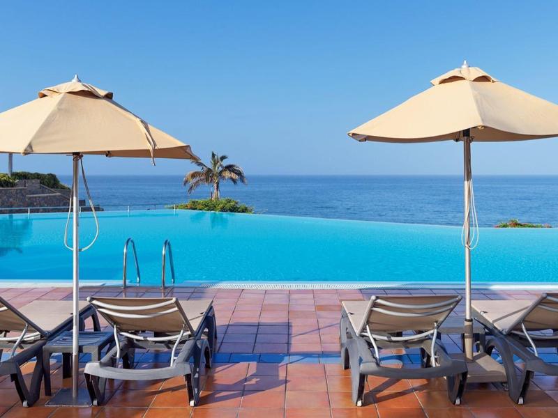 Einladender Infinity-Pool mit Liegestühlen und Sonnenschirmen, Meer und klarem Himmel im Hintergrund.