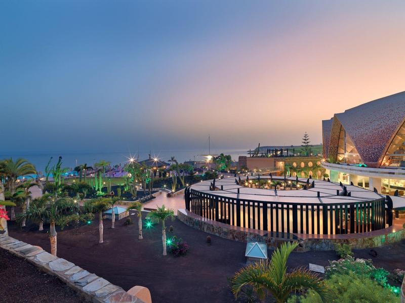 Landschaft mit moderner Architektur, beleuchteten Wegen, Pflanzen und Meer am Abend.