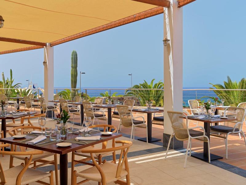 Terrassenrestaurant mit Holzstühlen, gedeckten Tischen und Blick auf das blaue Meer bei Sonnenschein.