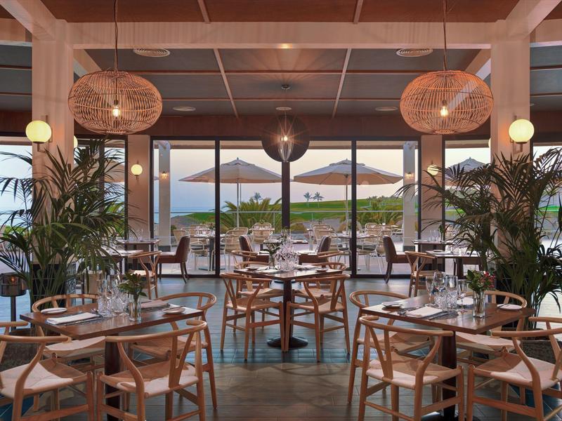 Modernes Restaurant mit Holztischen, Stühlen, Pflanzen und großem Fenster zum Außenbereich mit Meerblick.