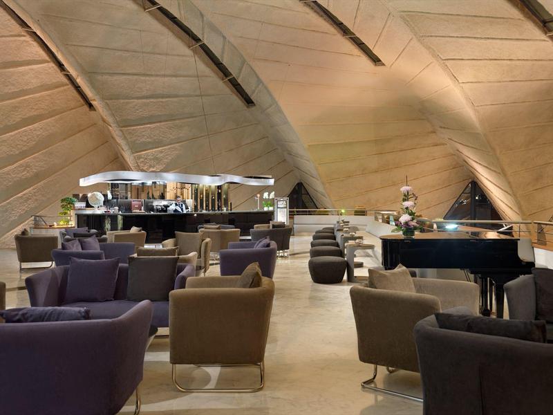 Moderner Loungebereich mit bequemen Sesseln, Bar im Hintergrund und Klavier unter gewölbter Decke.