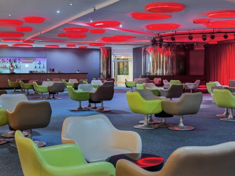 Großer moderner Veranstaltungsraum mit bunten Sesseln, roter Deckenbeleuchtung und Bar im Hintergrund.