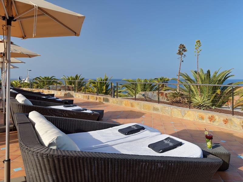 Mehrere Liegen mit weißen Polstern und schwarzen Handtüchern stehen unter Sonnenschirmen am Meer.