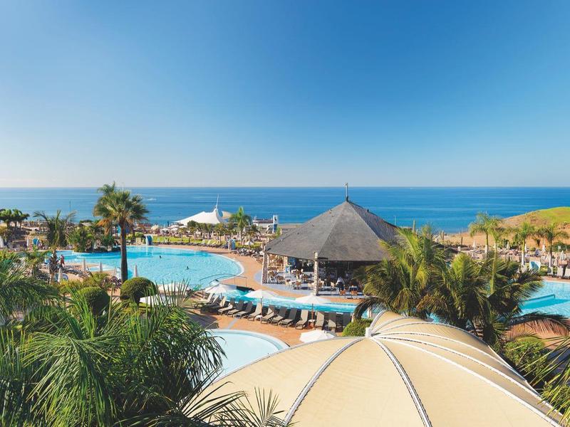 Badeanlage mit mehreren Pools, Palmen, Liegestühlen und Blick auf das blaue Meer bei klarem Himmel.