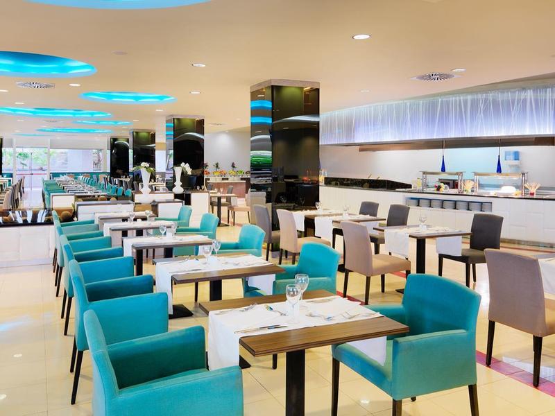 Moderner, heller Speisesaal mit türkisfarbenen Sesseln, weißen Tischen und großzügigem Buffetbereich.