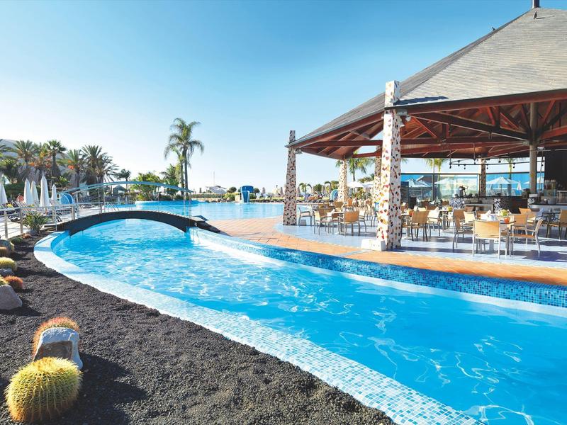 Moderner Außenpool mit blauem Wasser, Brücke, Palmen und überdachtem Sitzbereich bei klarem Himmel.