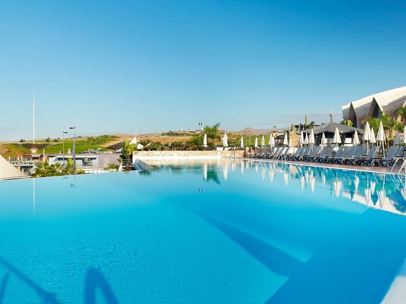 Großer Außenpool mit klar blauem Wasser, Sonnenschirmen und Palmen unter klarem Himmel.