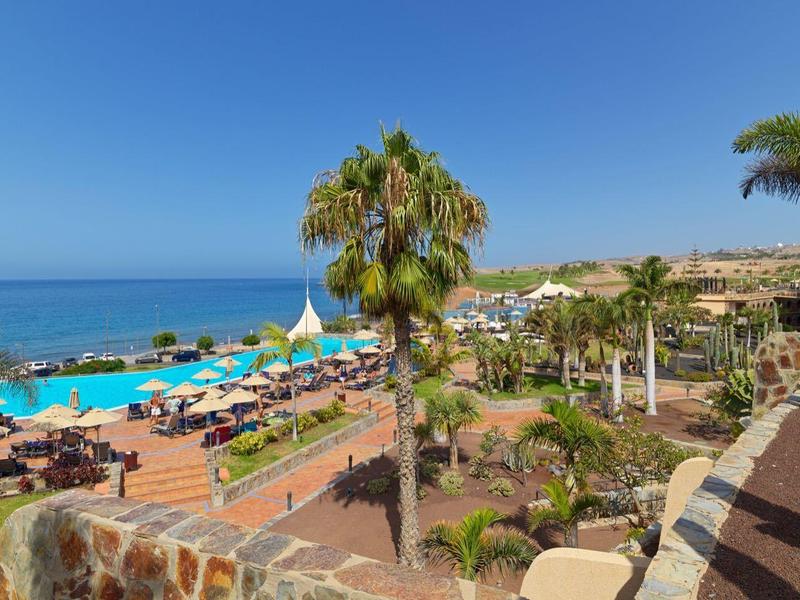 Strandpromenade mit Palmen, Sonne, blauem Meer und vielen Liegestühlen unter Sonnenschirmen.