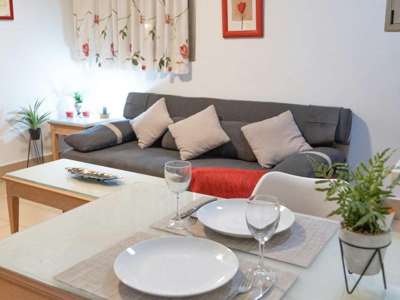 Przytulna jadalnia i salon z szarą sofą, jasnym stołem i dekoracyjnymi roślinami.