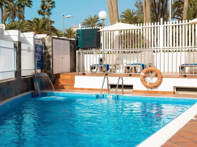 Mały basen z niebieskimi płytkami, otoczony palmami i białym ogrodzeniem.