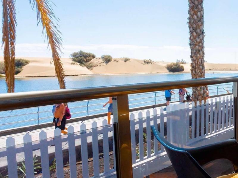 Widok na słoneczny taras z białymi ogrodzeniami, palmami i rzeką na pustyni.