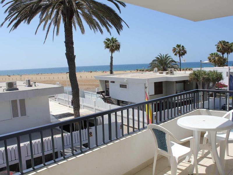 Balkon z stołem i krzesłami, widok na palmy, białe budynki i plażę w tle.