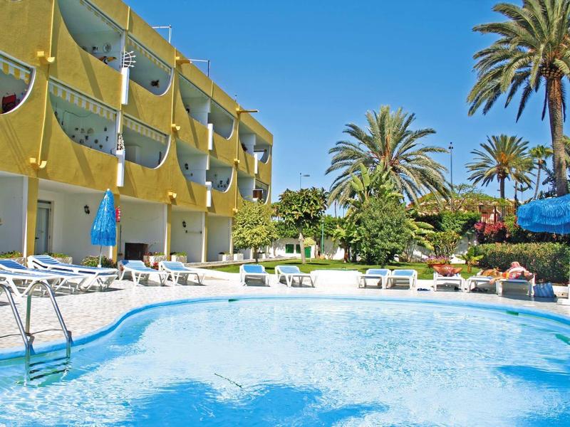 Hotel mit gelber Fassade, Balkon, Poolfläche und Liegestühlen unter blauem Himmel und Palmen.